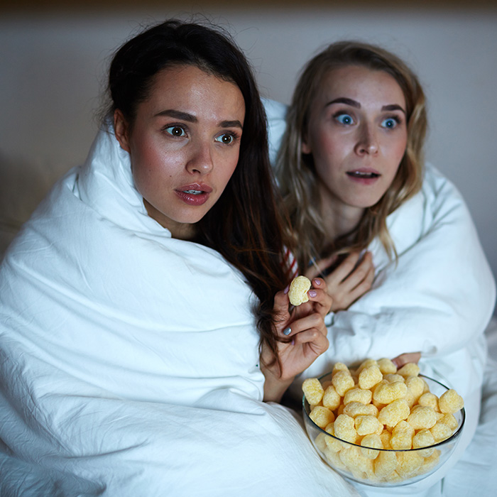 Two girls wrapped into blanket eating corn cheerios and watching horror movie